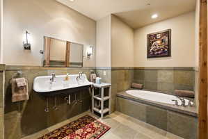 Bathroom featuring a wall-mounted trough sink with two faucets, a built-in soaking tub with a stone-finish deck, slate-style wall tiling, and coordinating floor tiles