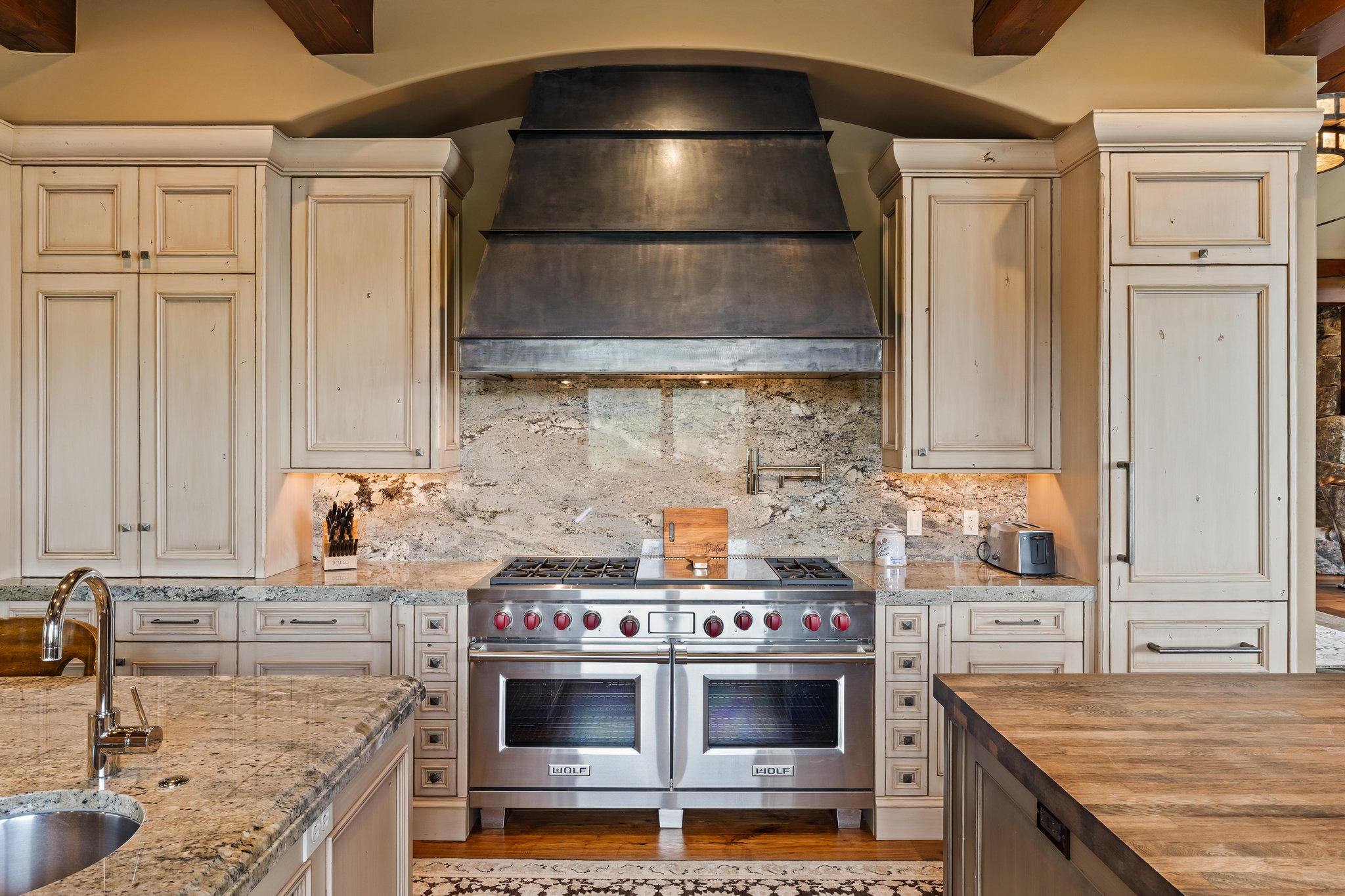 Gourmet kitchen featuring a professional-grade stainless steel range with double ovens, a matching custom metal range hood, stone slab countertops, a coordinating full-height stone backsplash, and custom wood cabinetry with distressed finish