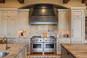 Gourmet kitchen featuring a professional-grade stainless steel range with double ovens, a matching custom metal range hood, stone slab countertops, a coordinating full-height stone backsplash, and custom wood cabinetry with distressed finish