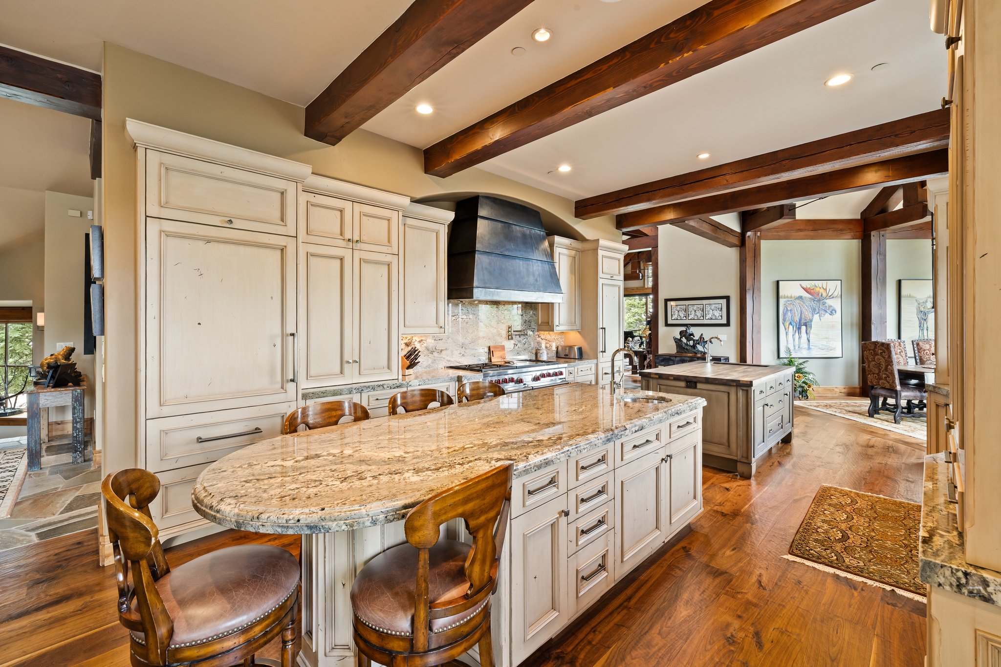 Gourmet kitchen featuring a large granite-topped island with seating, a built-in refrigerator, exposed ceiling beams, a custom range hood, and wood-finish flooring