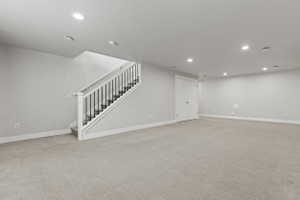 Spacious room featuring carpet flooring, recessed lighting, a staircase with white newel posts and black balusters, and a solid white door