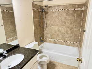 Bathroom featuring a built-in bathtub with a shower combination, neutral-toned wall tiles with a decorative mosaic accent band, a vanity with a dark speckled countertop, and a white integrated sink