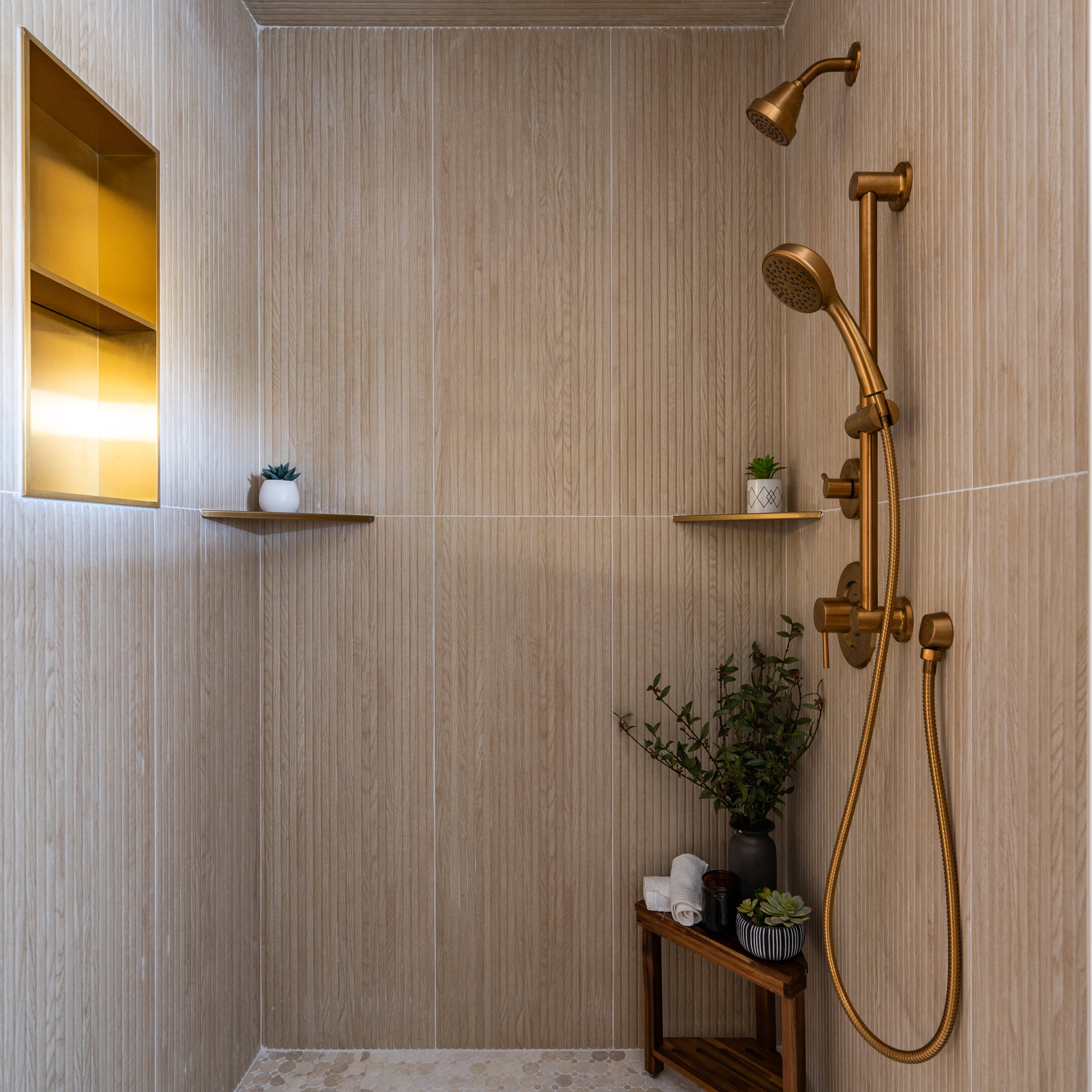 Shower stall featuring textured light-toned wall tile, a built-in brass-toned niche with integrated lighting, and a multi-function shower system with a handheld sprayer