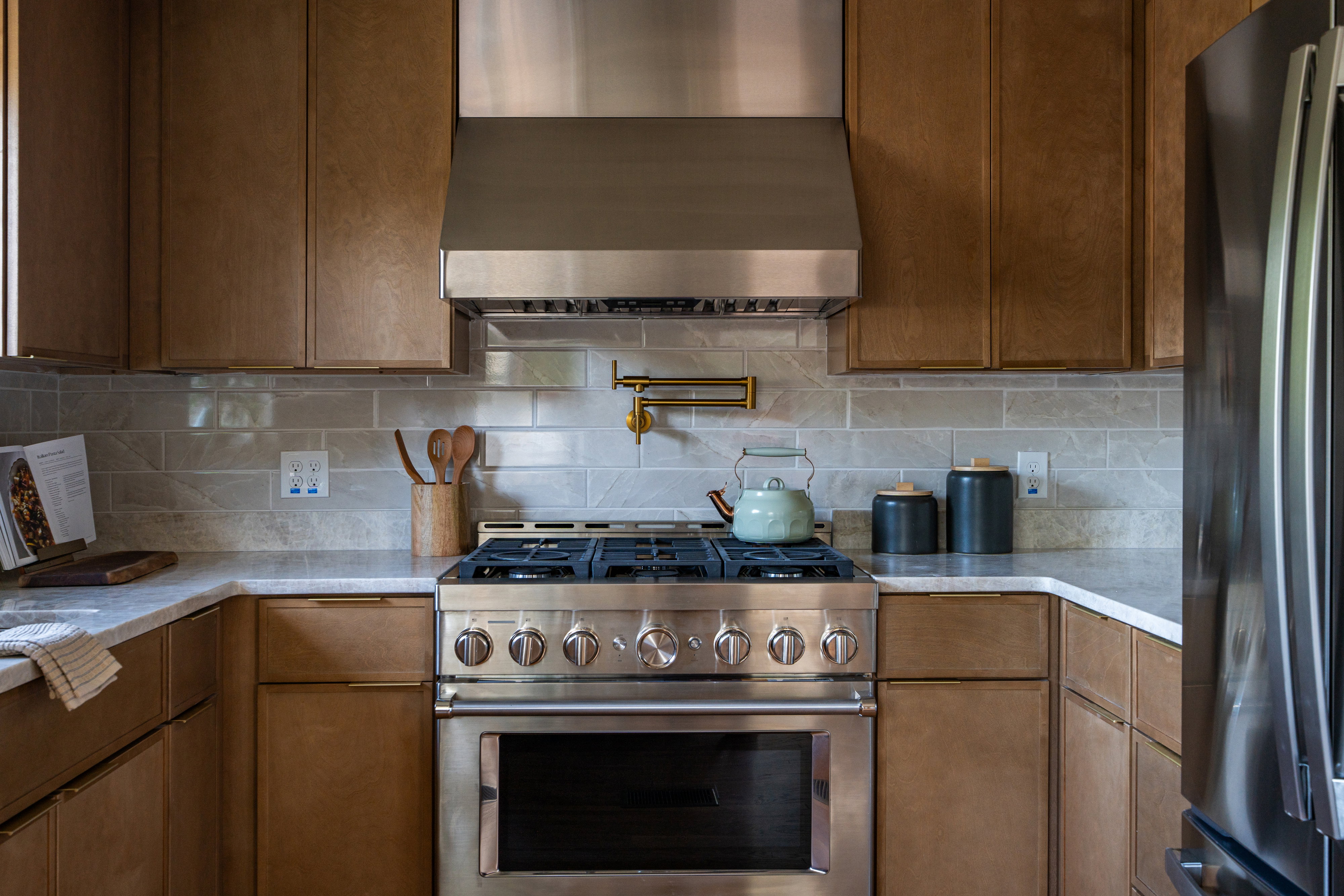 Gourmet kitchen featuring a stainless steel range with a pot filler, coordinating range hood, wood-finish cabinetry, and light-toned countertops with a subway tile backsplash