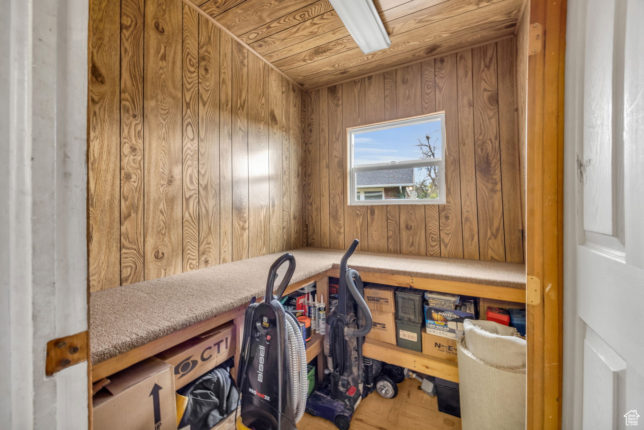 Utility room featuring wood-panel walls and ceiling, a single-pane window, integrated carpeted shelving, and a fluorescent ceiling light fixture