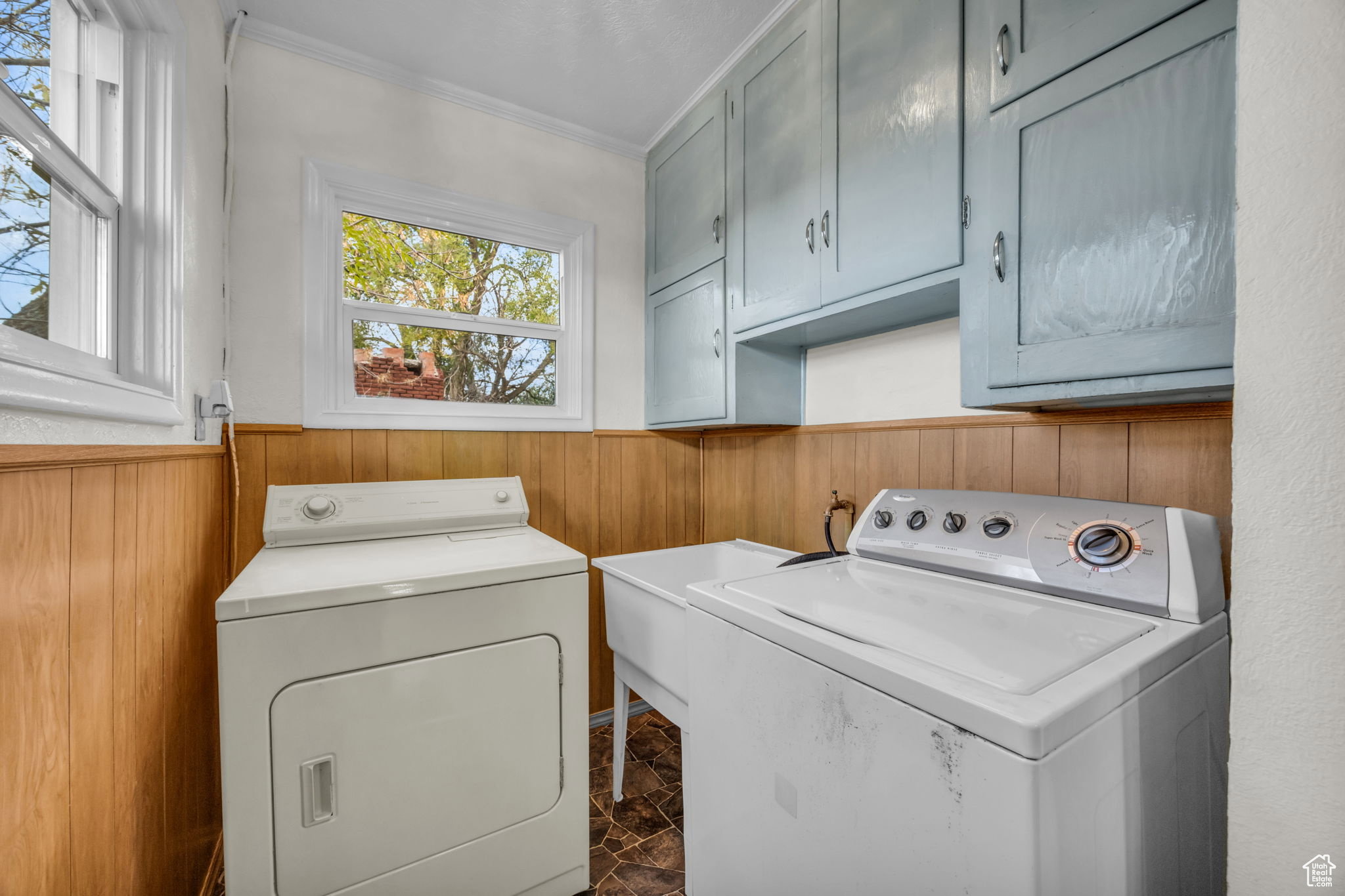 Utility room featuring wood paneling, upper cabinetry with light blue finish, a utility sink, and durable tile flooring