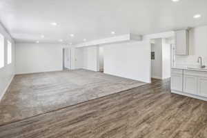 Spacious room featuring wood-finish flooring transitioning to neutral-toned carpeting, recessed lighting, and a kitchenette area with light gray cabinetry, a countertop, and a stainless steel sink with a gooseneck faucet