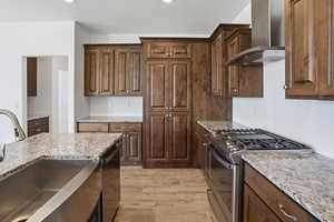 Kitchen featuring wood-finish cabinetry, light-toned granite countertops, a stainless steel farmhouse sink, a stainless steel gas range with a matching range hood, and wood-finish flooring