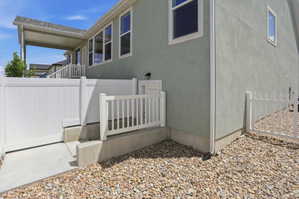Rear exterior of the property featuring a covered patio, light green stucco siding, white vinyl fencing, and a gravel-covered ground