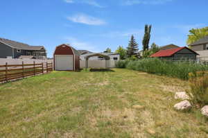 Expansive grassy yard featuring a red barn-style shed with a white garage door, a metal carport, and a rustic wood fence