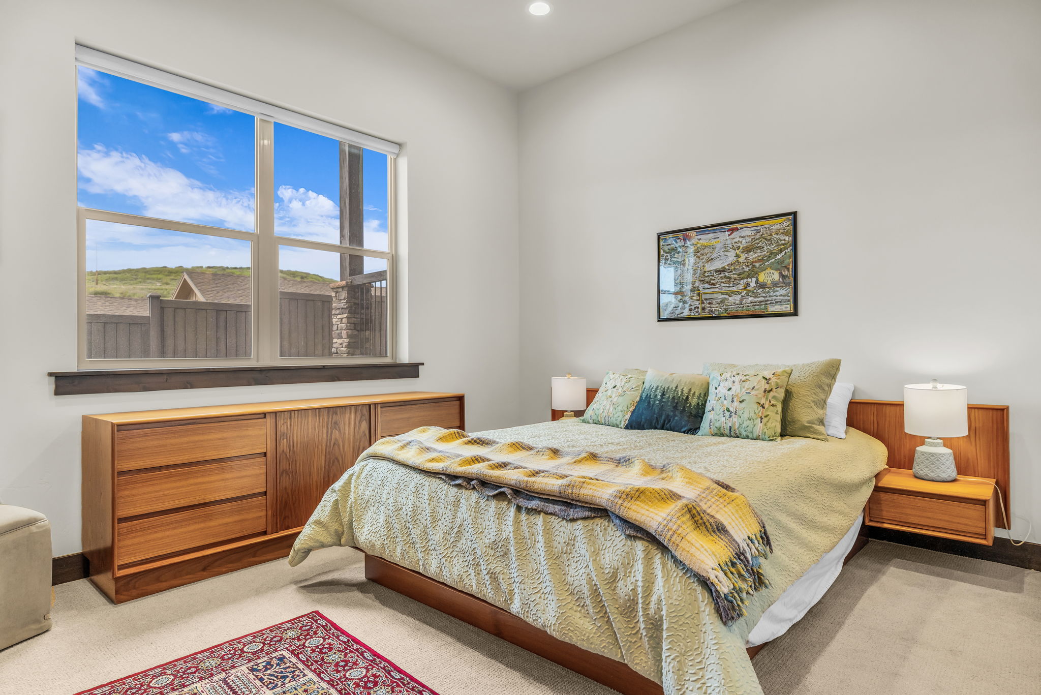 Spacious bedroom with a large window, built-in wooden headboard, integrated bedside tables, and light carpet flooring