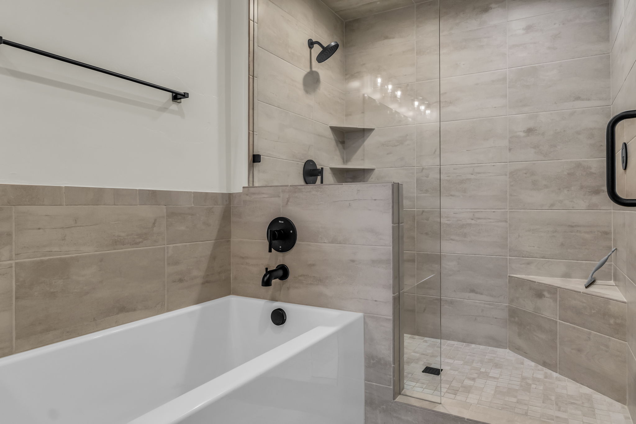Modern bathroom featuring a soaking tub, glass-enclosed walk-in shower, dark-finish fixtures, light-toned tile surrounds, and integrated shower seating