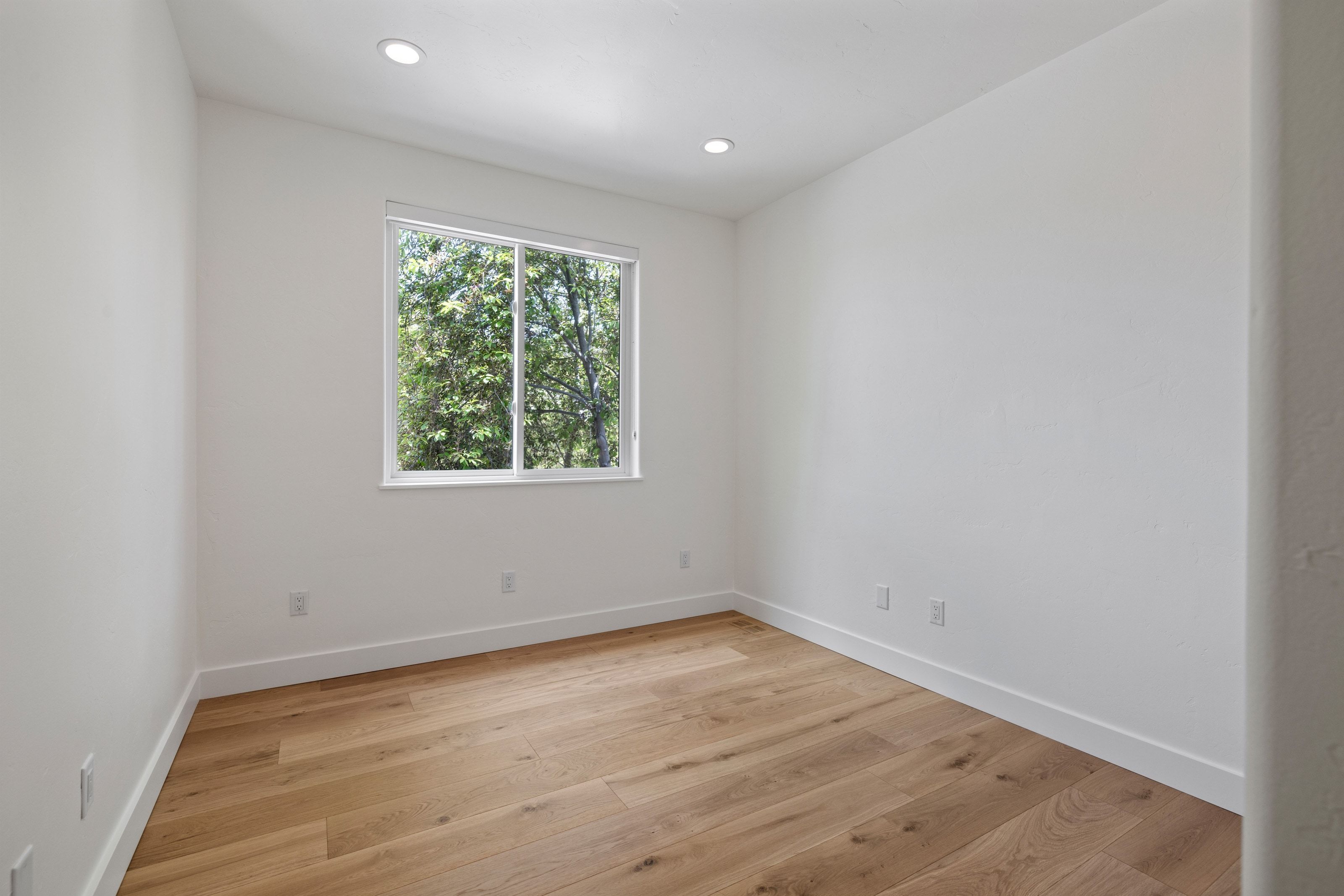 Room featuring wood-finish flooring, white walls, recessed lighting, and a window with a green tree backdrop