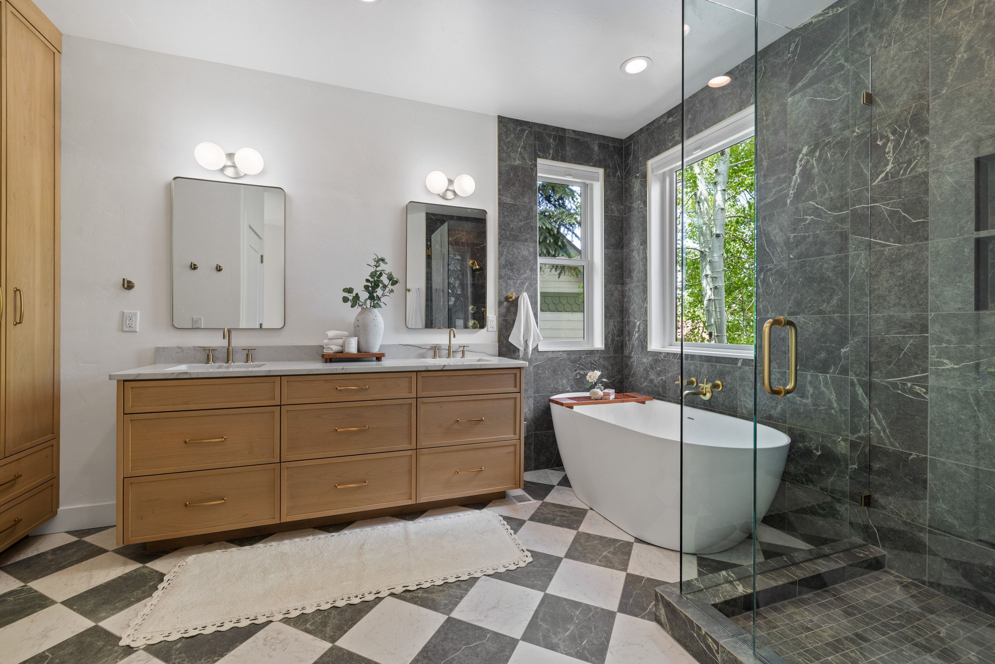 Spacious bathroom featuring a freestanding soaking tub, a large glass-enclosed shower with dark stone-finish tile, a double vanity with stone countertops and brass fixtures, and checkered tile flooring