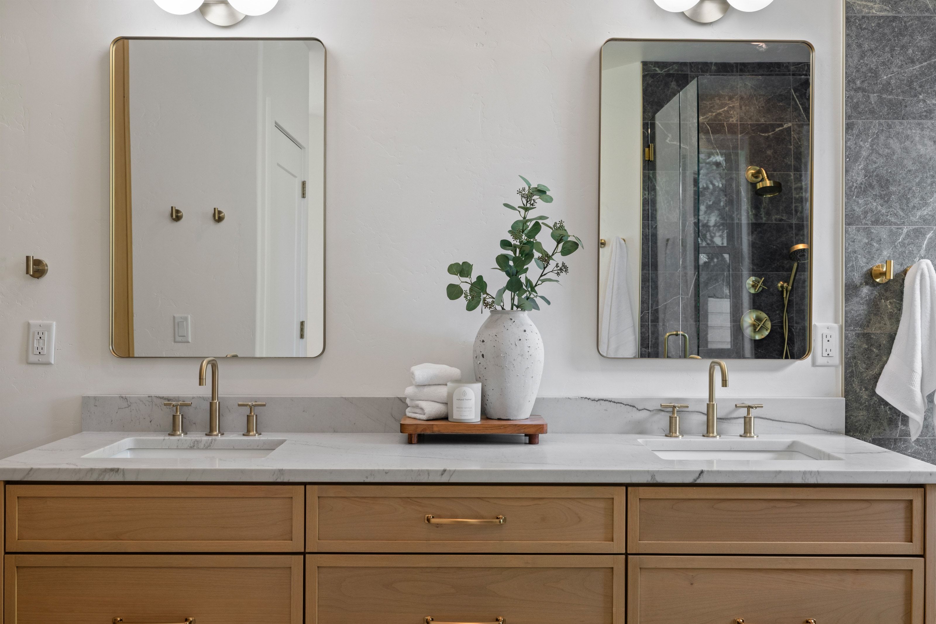Dual vanity featuring light wood cabinetry, white stone countertops, integrated sinks, and individual gold-framed mirrors