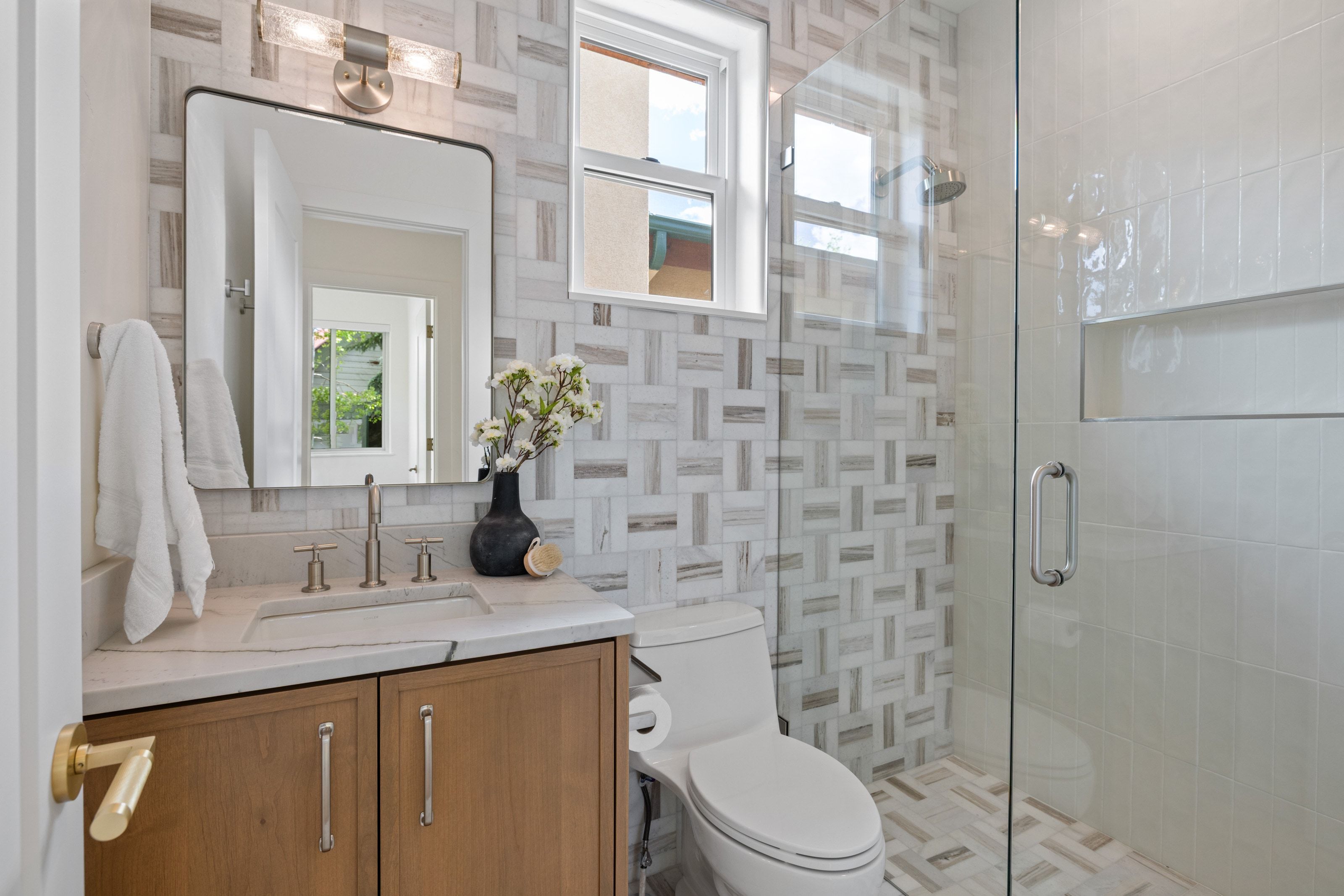 Contemporary bathroom featuring a frameless glass shower enclosure, a wood-finish vanity with a light-toned countertop, a rectangular sink with brushed nickel fixtures, a framed mirror, and patterned wall tiling