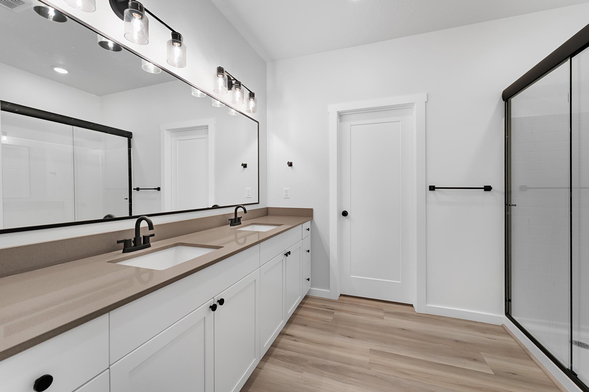 Bathroom featuring a dual vanity with a tan countertop, wood-finish flooring, a frameless glass shower enclosure, and matte black hardware throughout