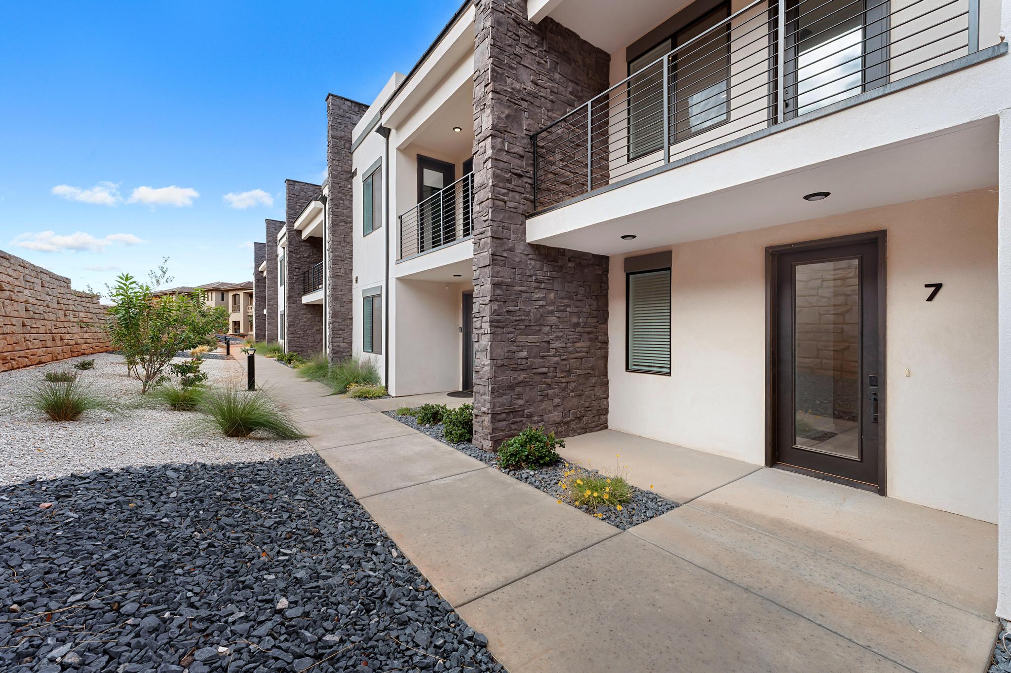 Modern multi-level building exterior featuring stacked stone accents, stucco siding, and a private balcony with horizontal metal railings