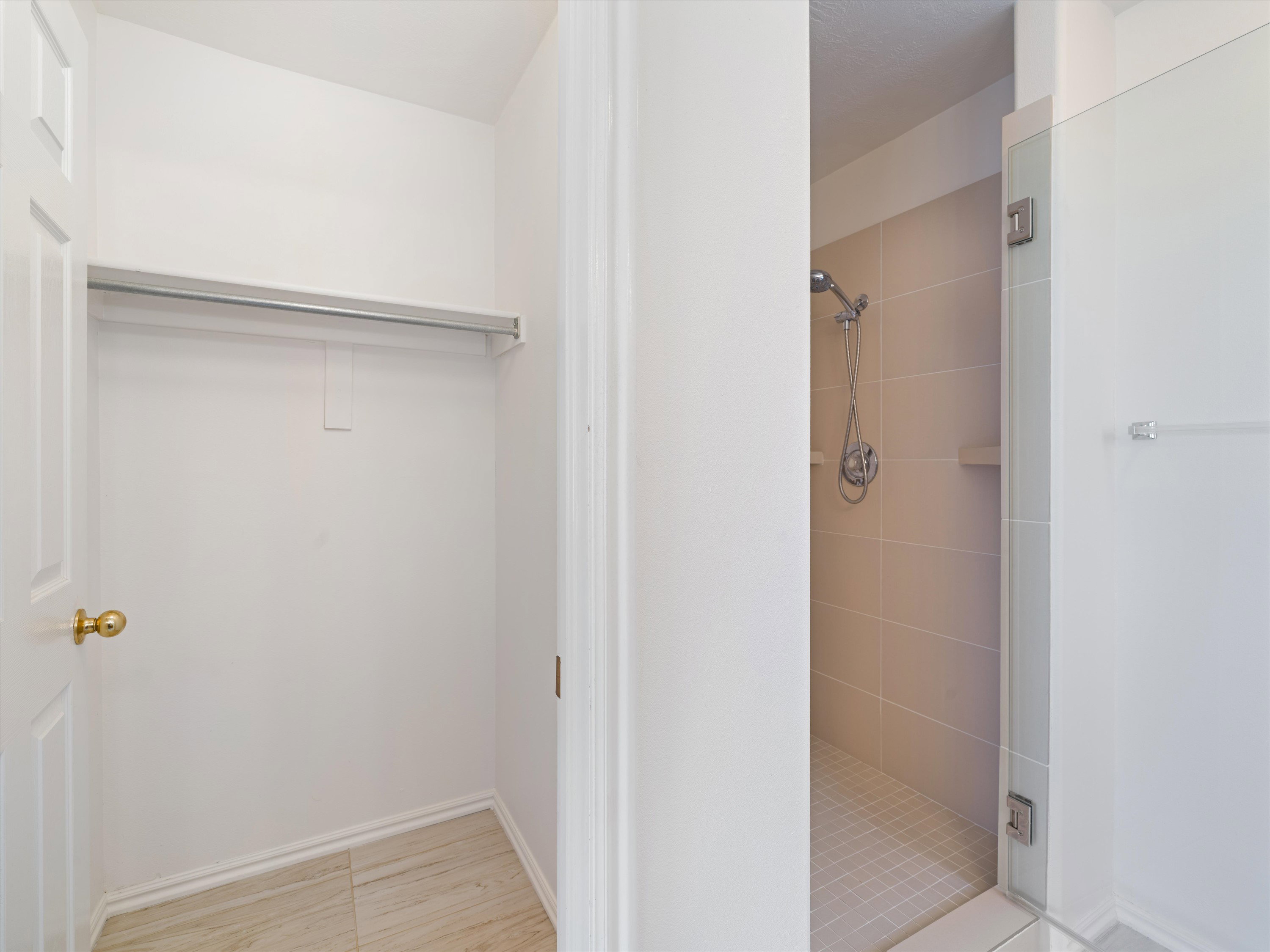 Recessed closet space with a clothing rod and light-toned tile flooring, complemented by a nearby tiled shower enclosure with a handheld showerhead and built-in shelf