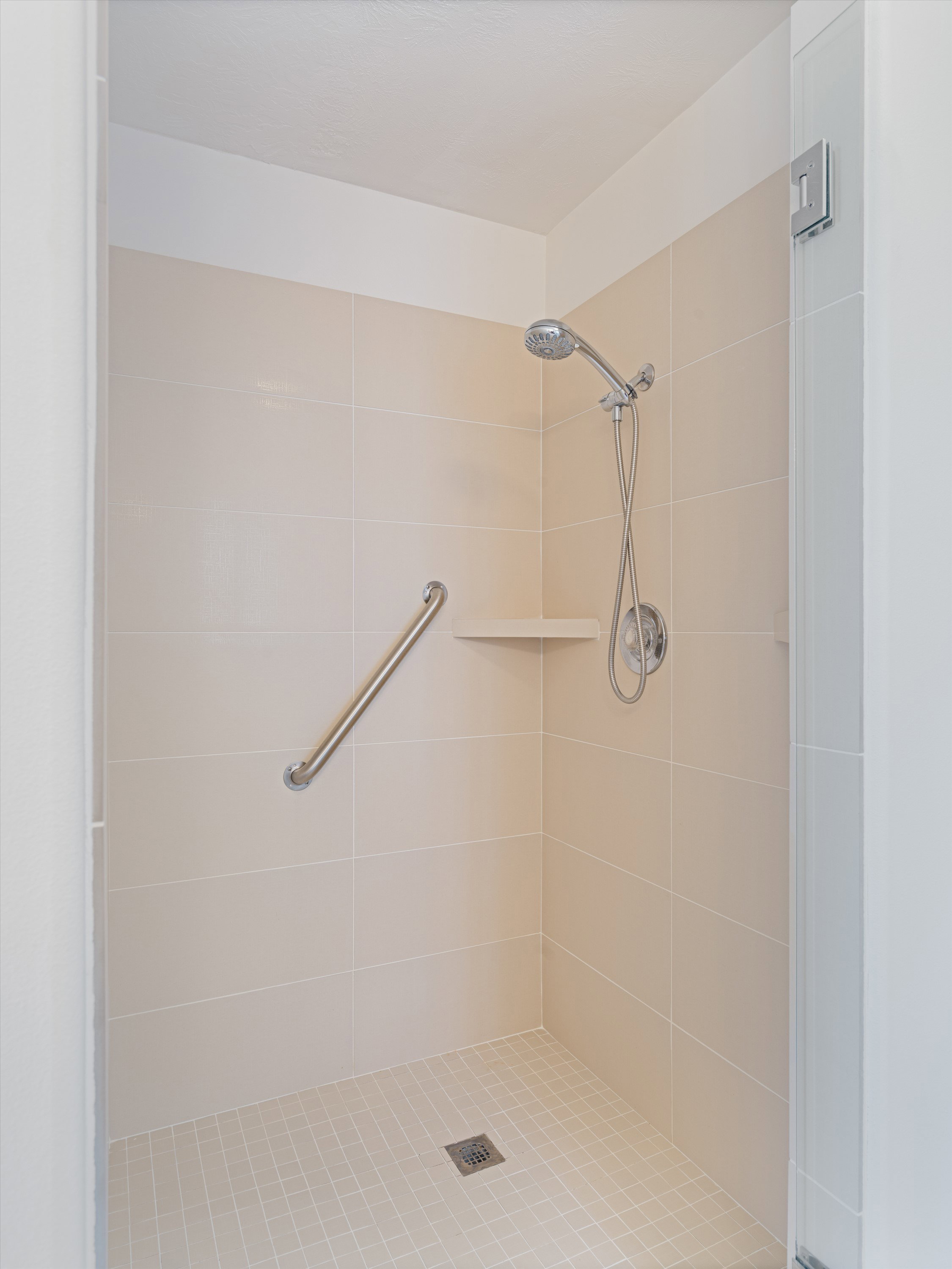 Walk-in shower with neutral wall tiling, a handheld showerhead, corner shelf, and grab bar