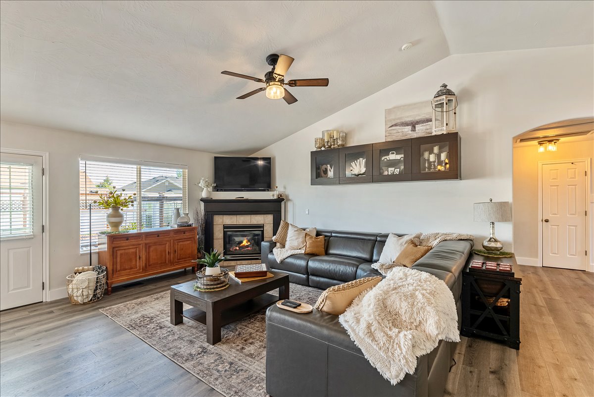 Vaulted ceiling living area featuring wood-finish flooring, a fireplace with a tile surround and dark mantle, a ceiling fan, and a window with blinds