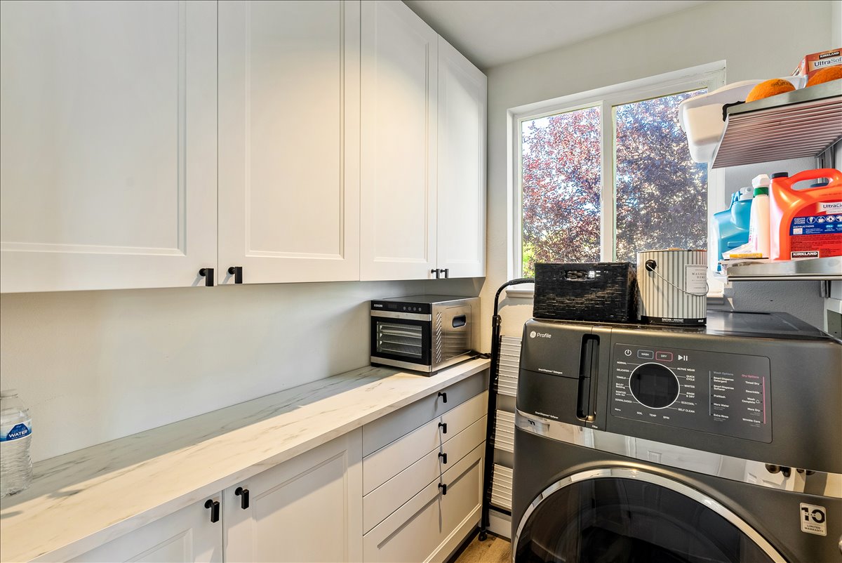 Contemporary laundry area featuring white shaker-style cabinetry, a light-toned countertop, a window with exterior foliage, and recessed lighting