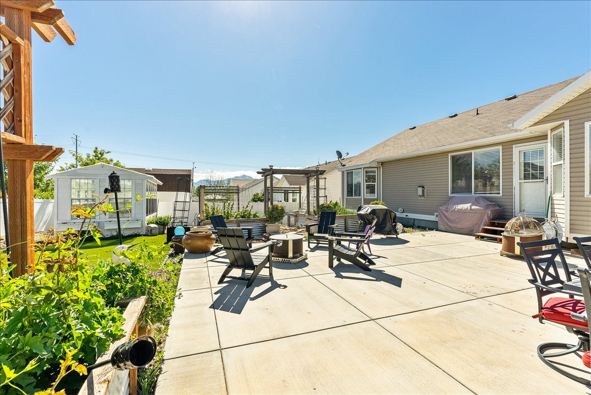 Expansive concrete patio with a fire pit area, surrounded by a manicured lawn, raised garden beds, and a white greenhouse structure