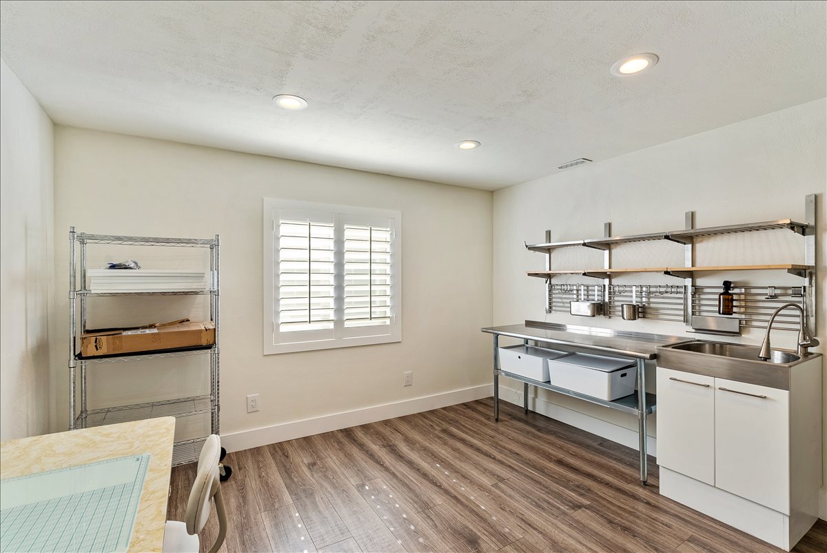 Versatile workspace featuring wood-finish flooring, a stainless steel workstation with integrated sink, white cabinetry, recessed lighting, and a window with white louvered shutters