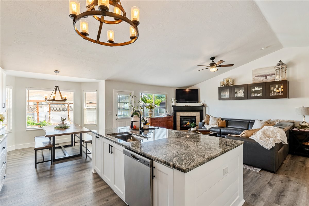 Spacious open-concept interior featuring a substantial kitchen island with a stone countertop and built-in sink, wood-finish flooring, white cabinetry with black hardware, a dining nook with a bay window, and a living area with a fireplace