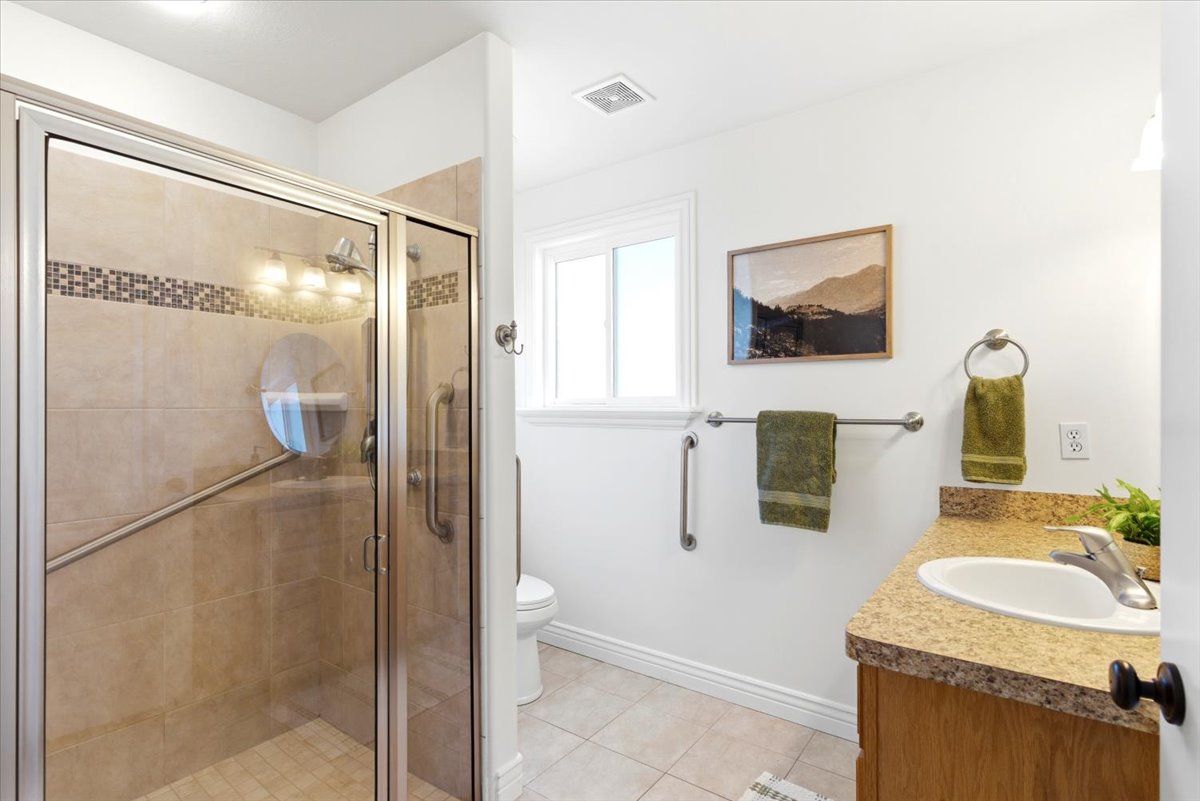 Glass-enclosed shower featuring neutral-toned tile work with mosaic accent banding, a window providing natural light, a wood-finish vanity with a speckled countertop and integrated sink, and tile flooring