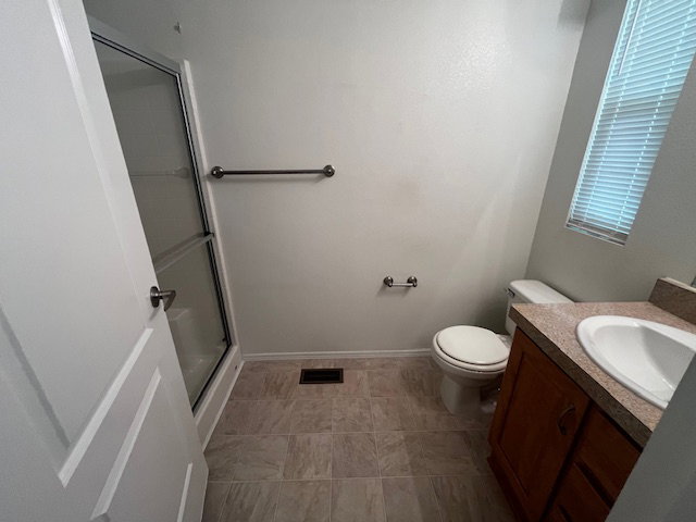Shower with sliding glass enclosure, tile flooring, window with blinds, wood-finish vanity with integrated sink, and toilet