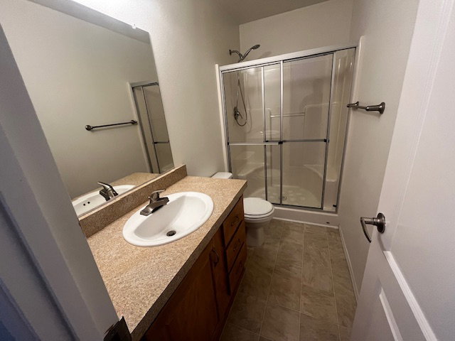 Bathroom featuring a shower with sliding glass doors, a vanity with a drop-in sink, a speckled countertop, and wood-finish cabinetry, all complemented by light-toned flooring