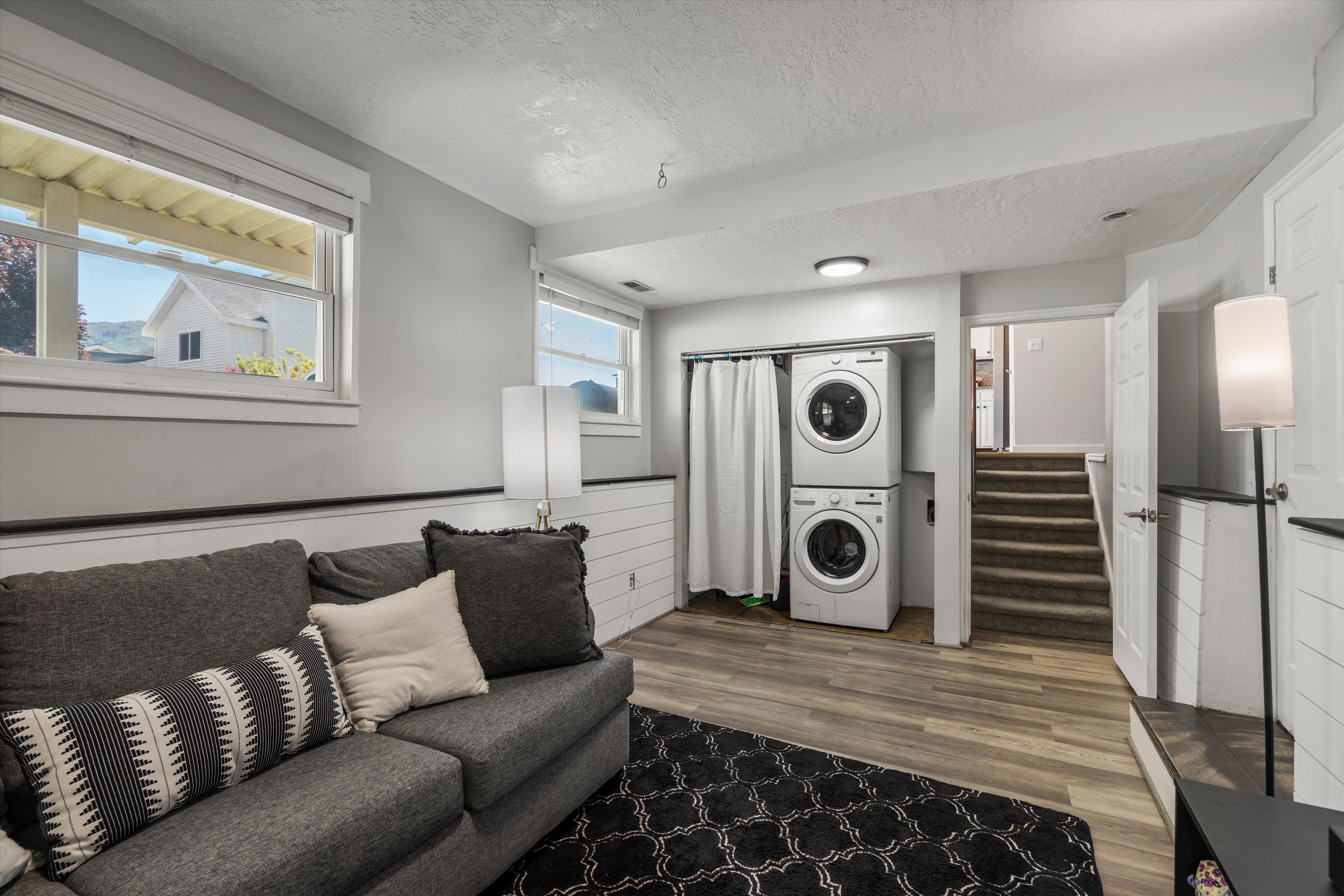 Lower-level living area featuring wood-finish flooring, two windows with blinds, and white painted walls with a shiplap accent