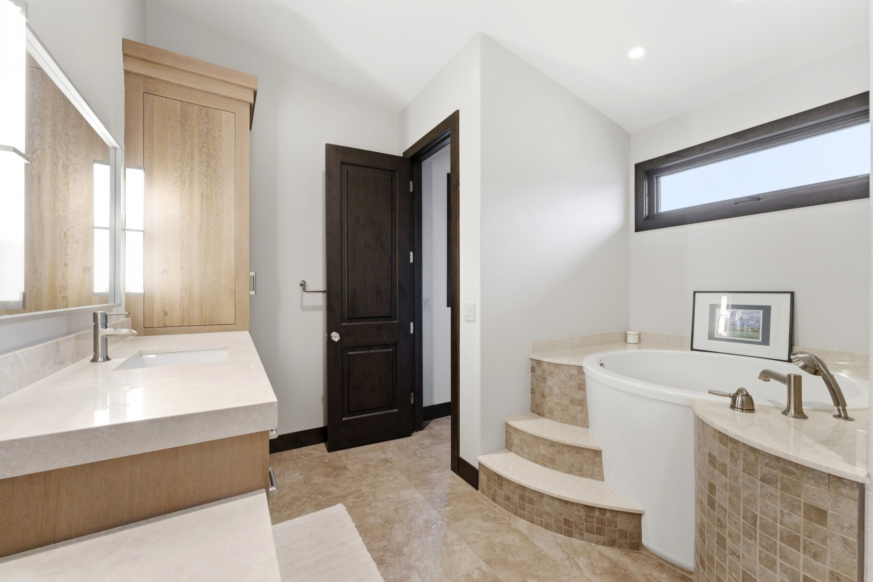 Contemporary bathroom featuring a built-in soaking tub with mosaic tile surround, a light wood-finish vanity with an integrated sink, a large mirror with integrated lighting, and tile flooring