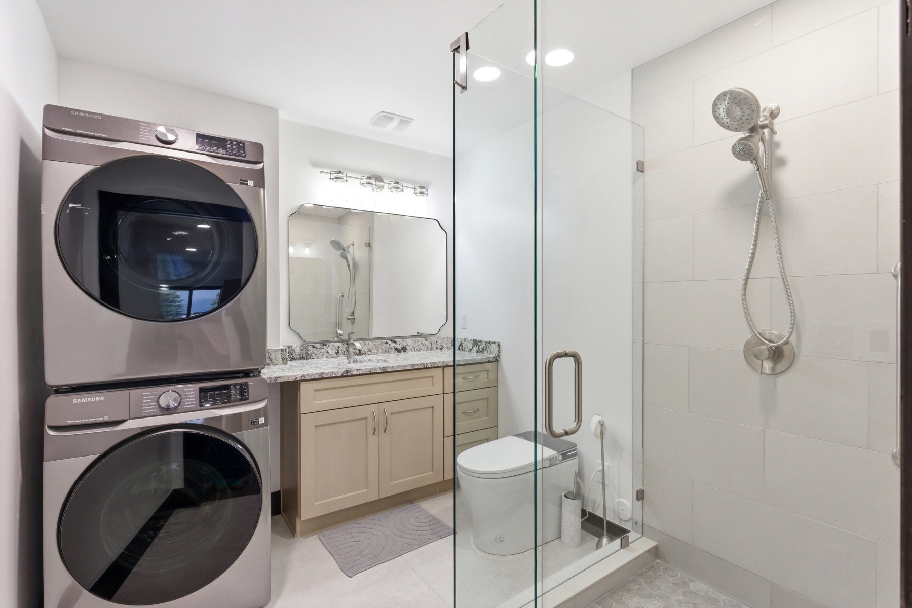 Modern bathroom featuring a stacked laundry unit, a glass-enclosed shower with a handheld fixture, light wood-finish vanity with a stone countertop, and a decorative wall mirror