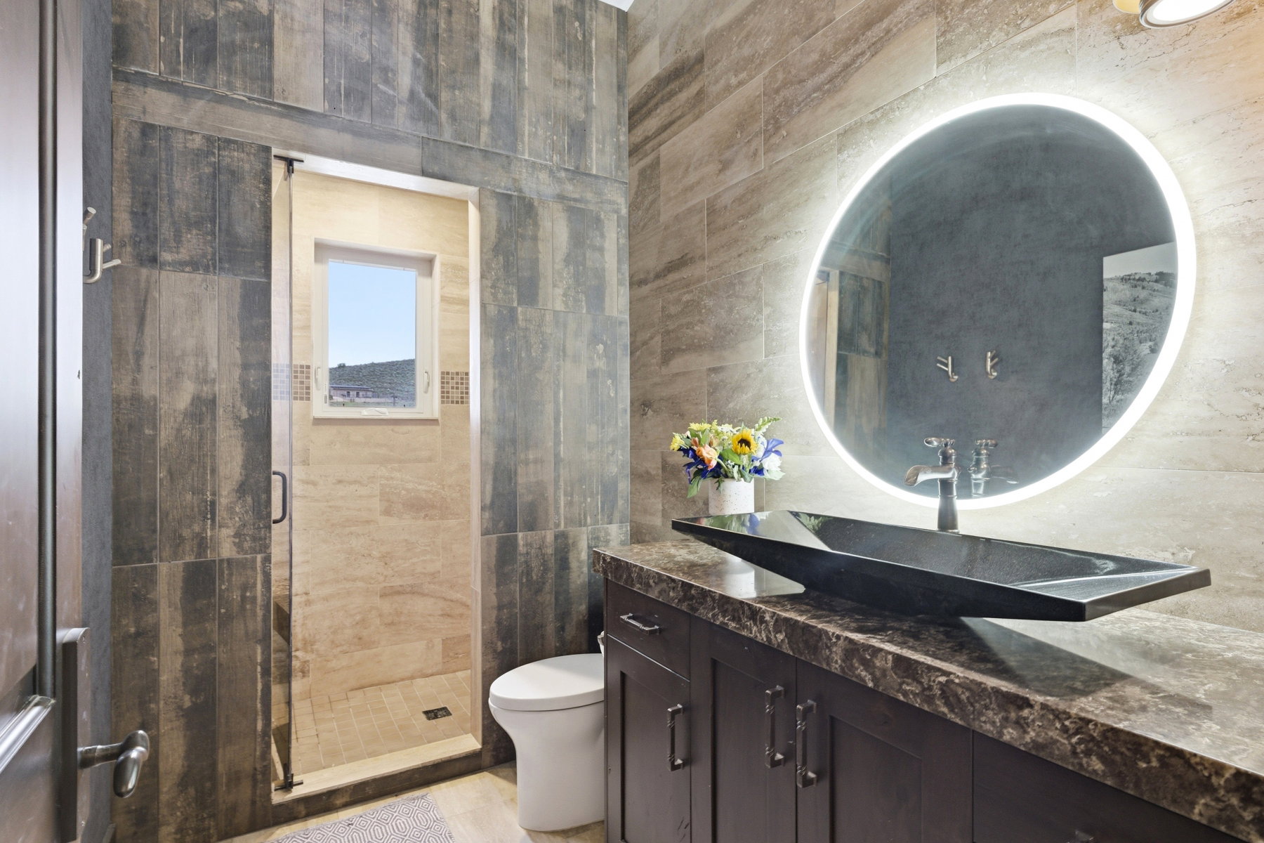 Bathroom featuring a contemporary vanity with an integrated vessel sink, an illuminated mirror, and a walk-in shower with natural stone-look tiling