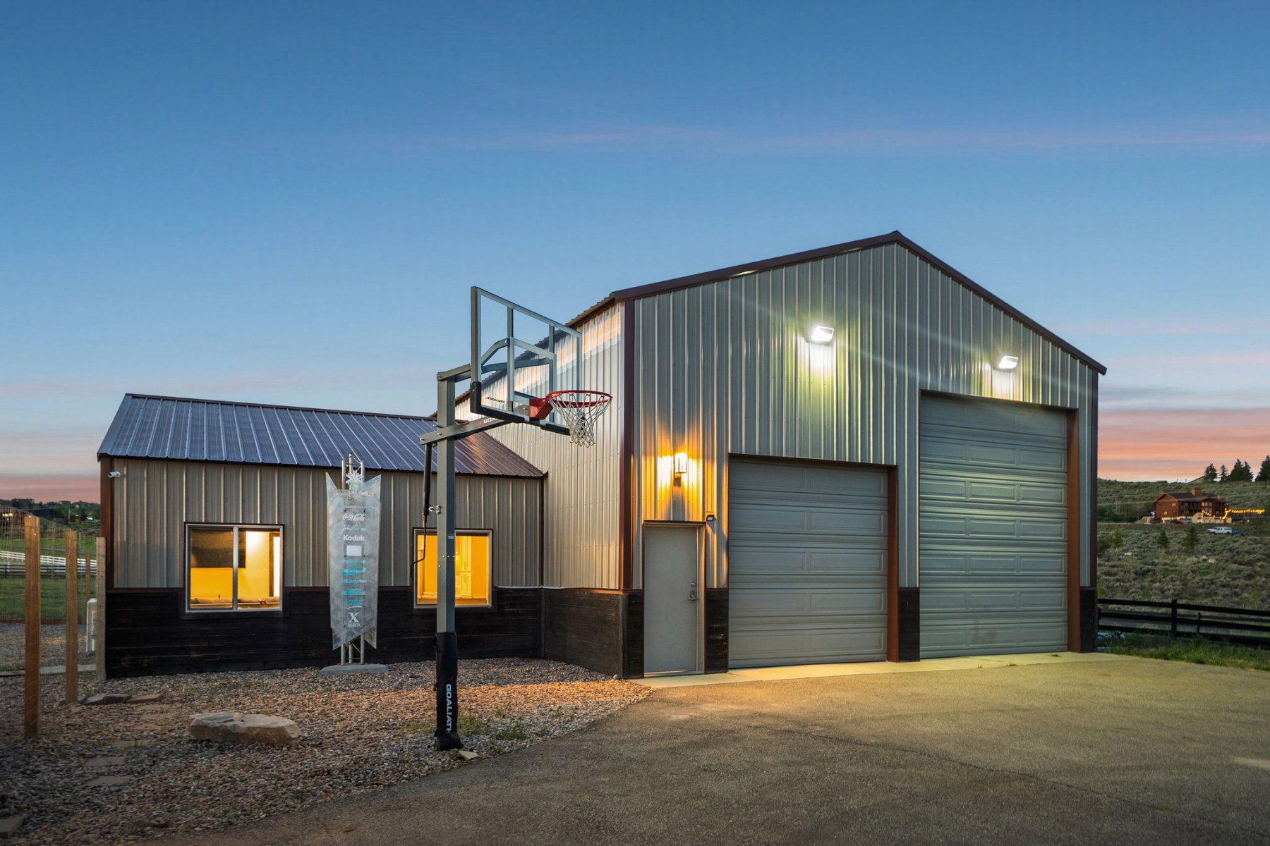 Metal-sided structure featuring two roll-up garage doors, a side entry door, and a built-in basketball hoop