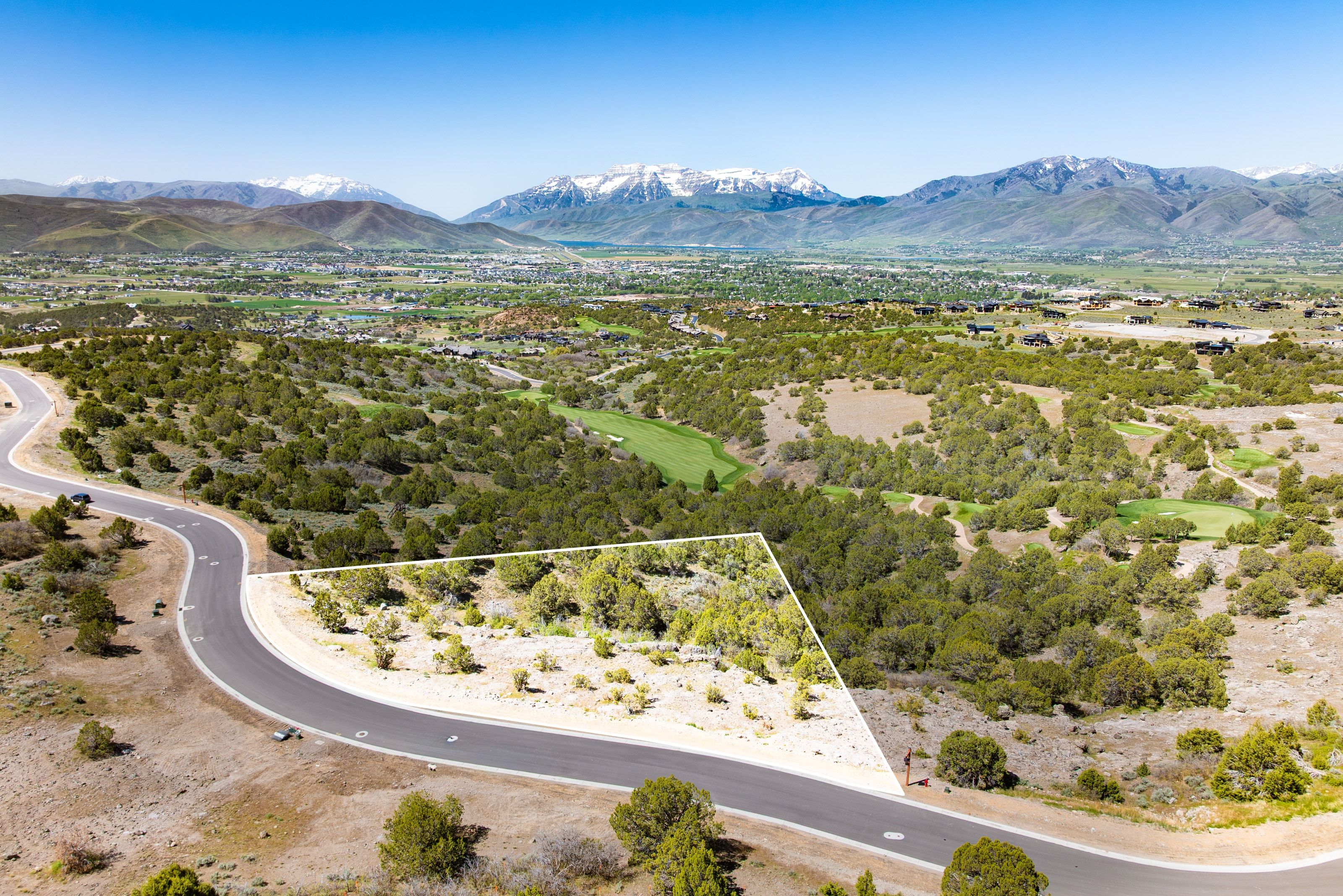 Aerial perspective of a vacant lot outlined in white, featuring natural vegetation and a paved road frontage