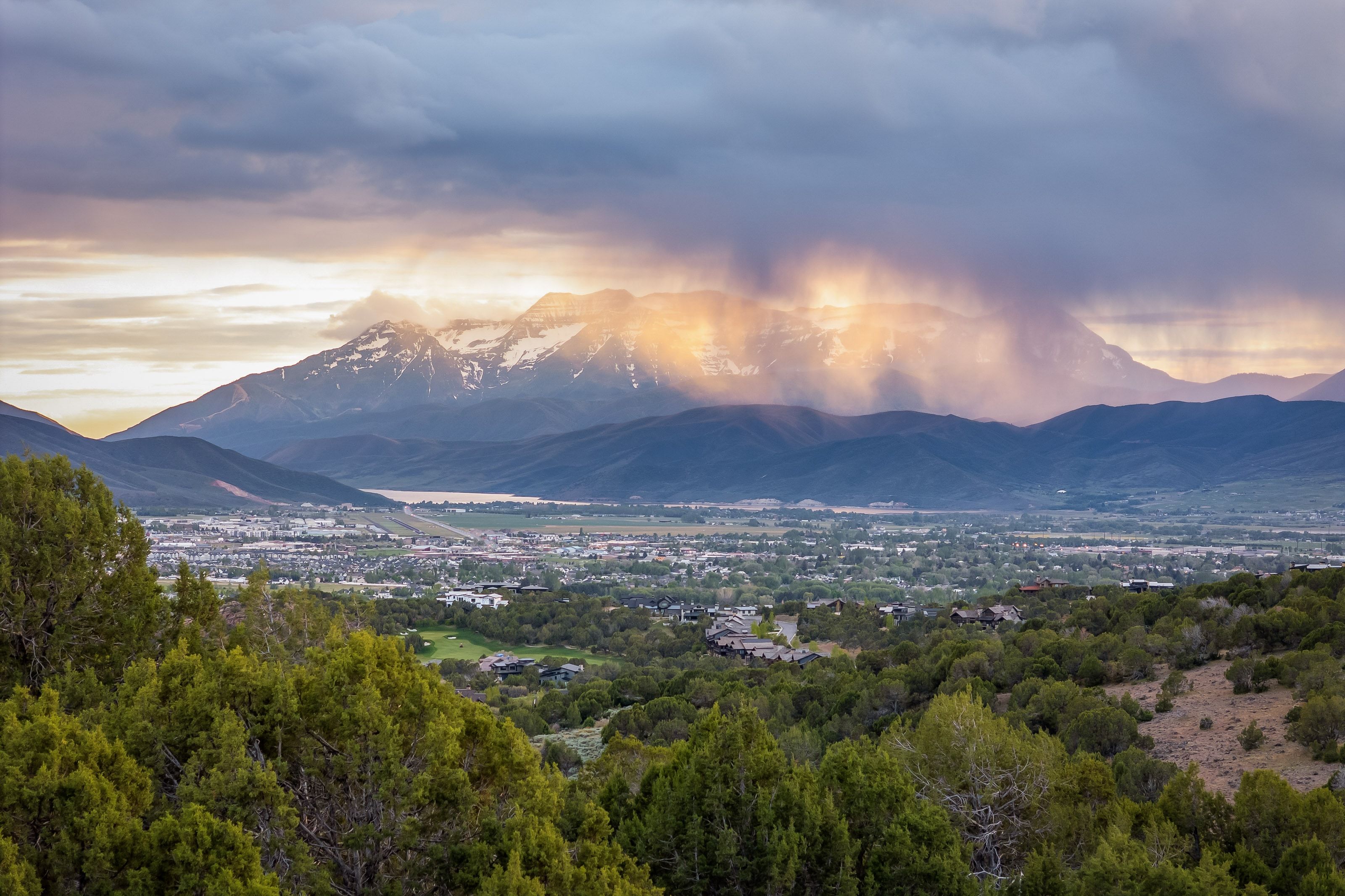 Expansive mountain and valley scenery featuring distant snow-capped peaks, a vast valley floor with a townscape, and a body of water
