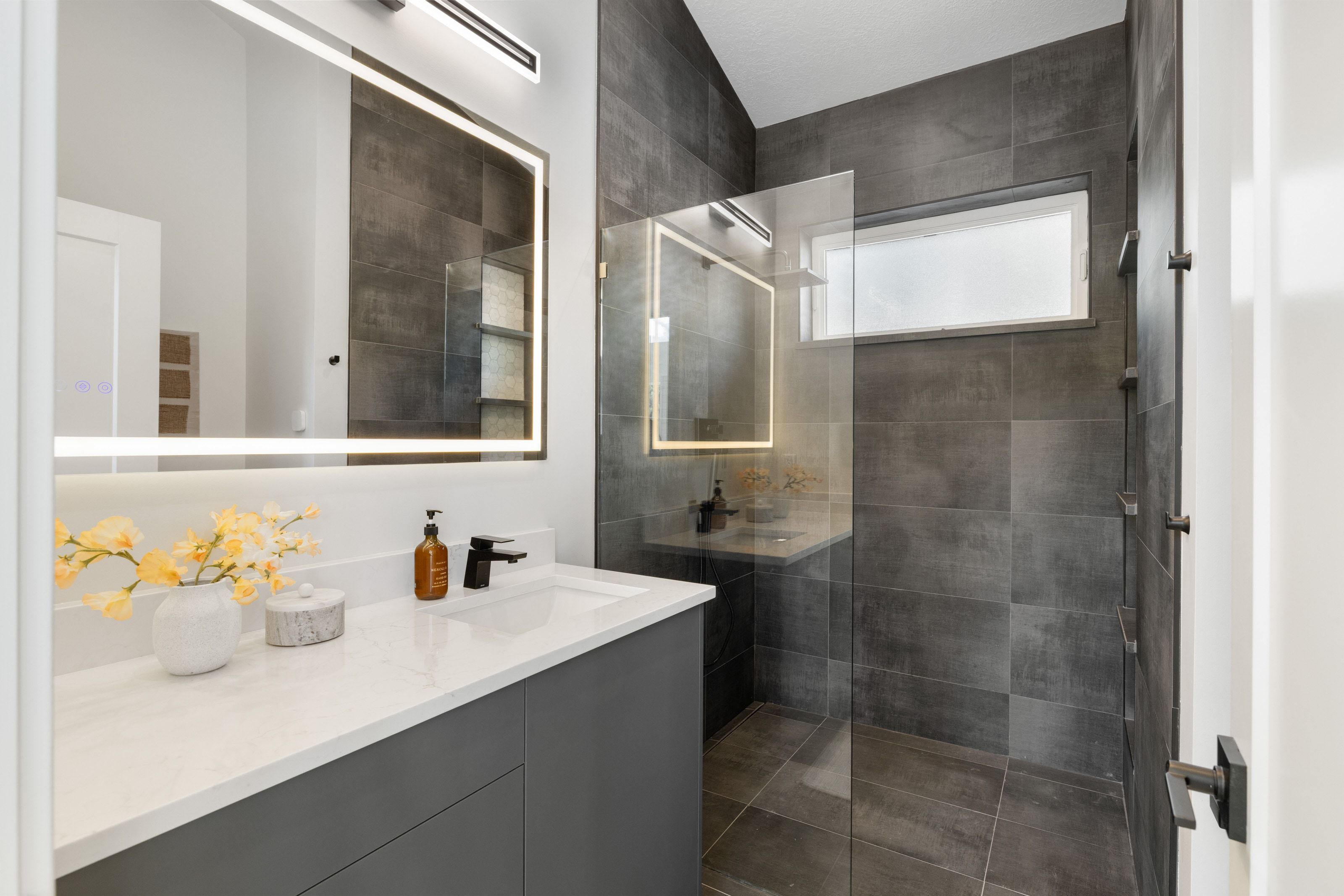 Contemporary bathroom featuring a grey vanity with a white countertop, an illuminated mirror, and a walk-in shower with dark grey tile surrounds and a glass enclosure