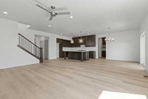 Spacious open-concept living area featuring light wood-finish flooring, a contemporary kitchen with dark wood cabinetry and a central island, a modern chandelier, and a staircase with dark wood handrails and black balusters
