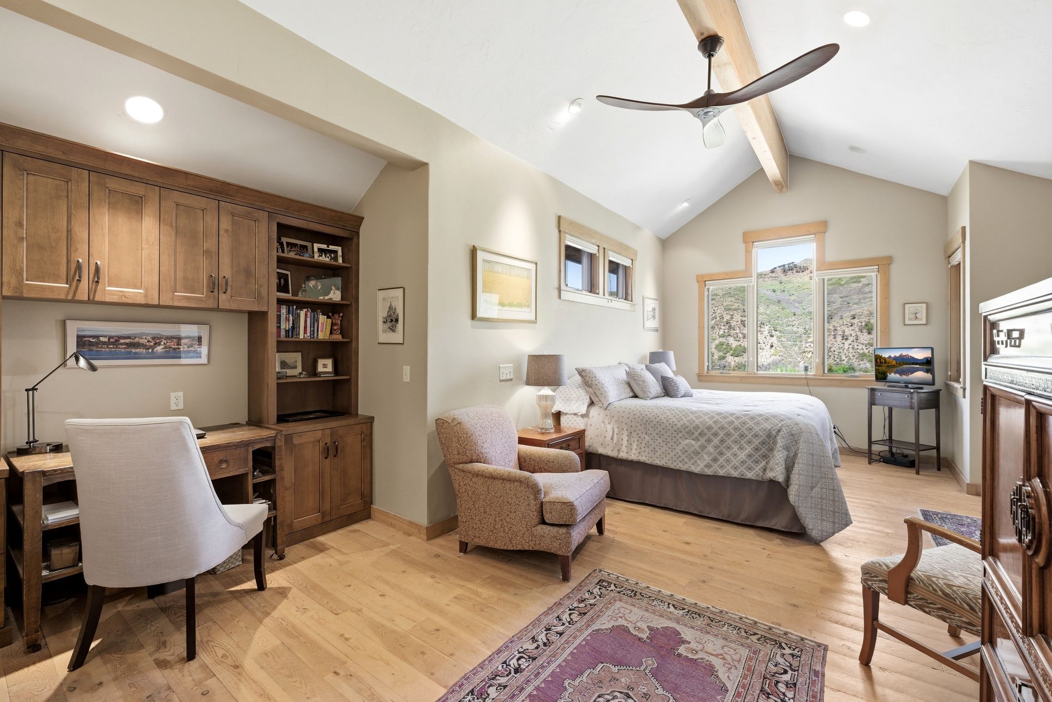 Vaulted ceiling with exposed wood beam, natural wood-finish flooring, built-in cabinetry with desk, and multiple windows offering hillside scenery