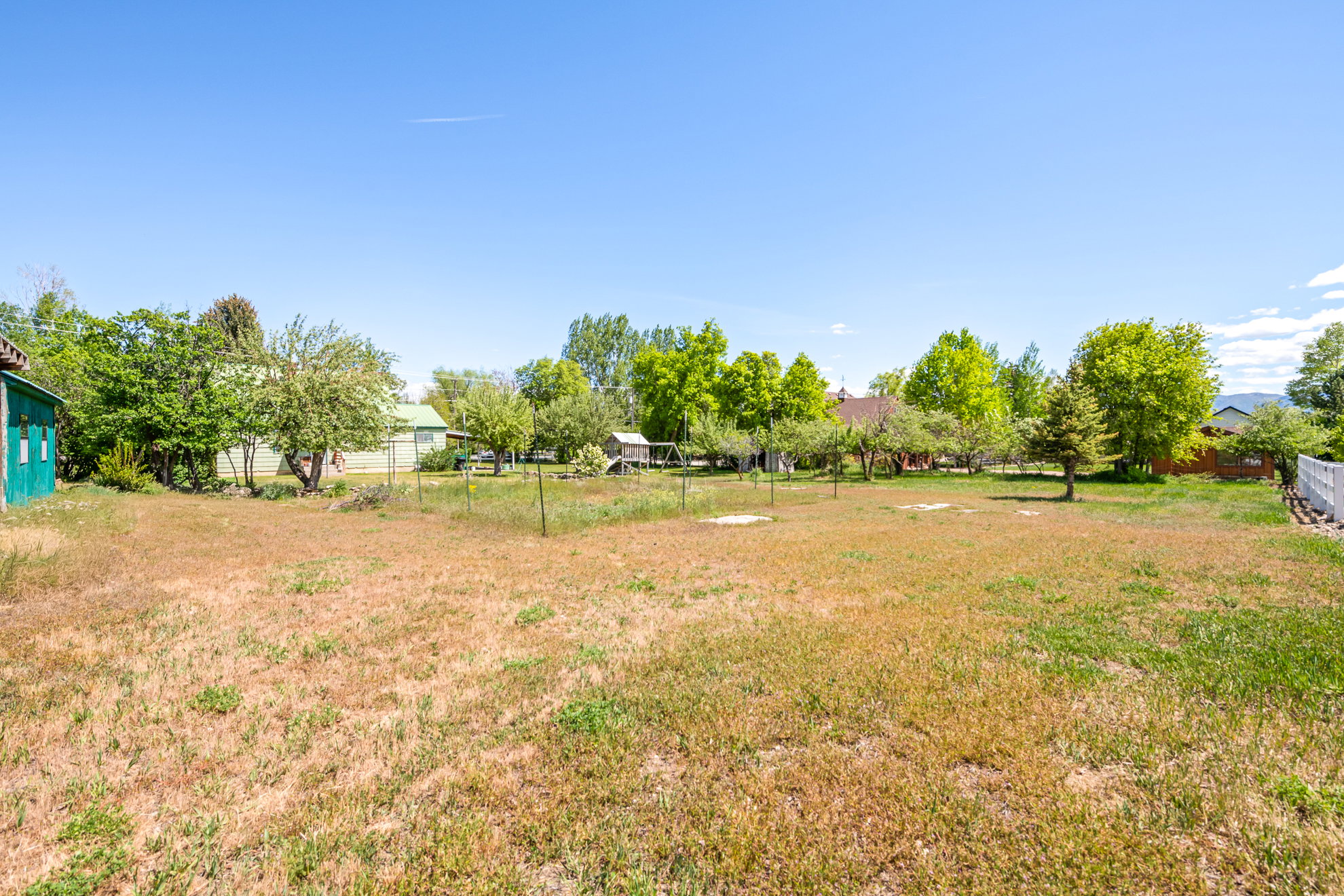 Expansive open lot featuring a clear blue sky, numerous mature trees, a partial white fence, and a distant mountain range