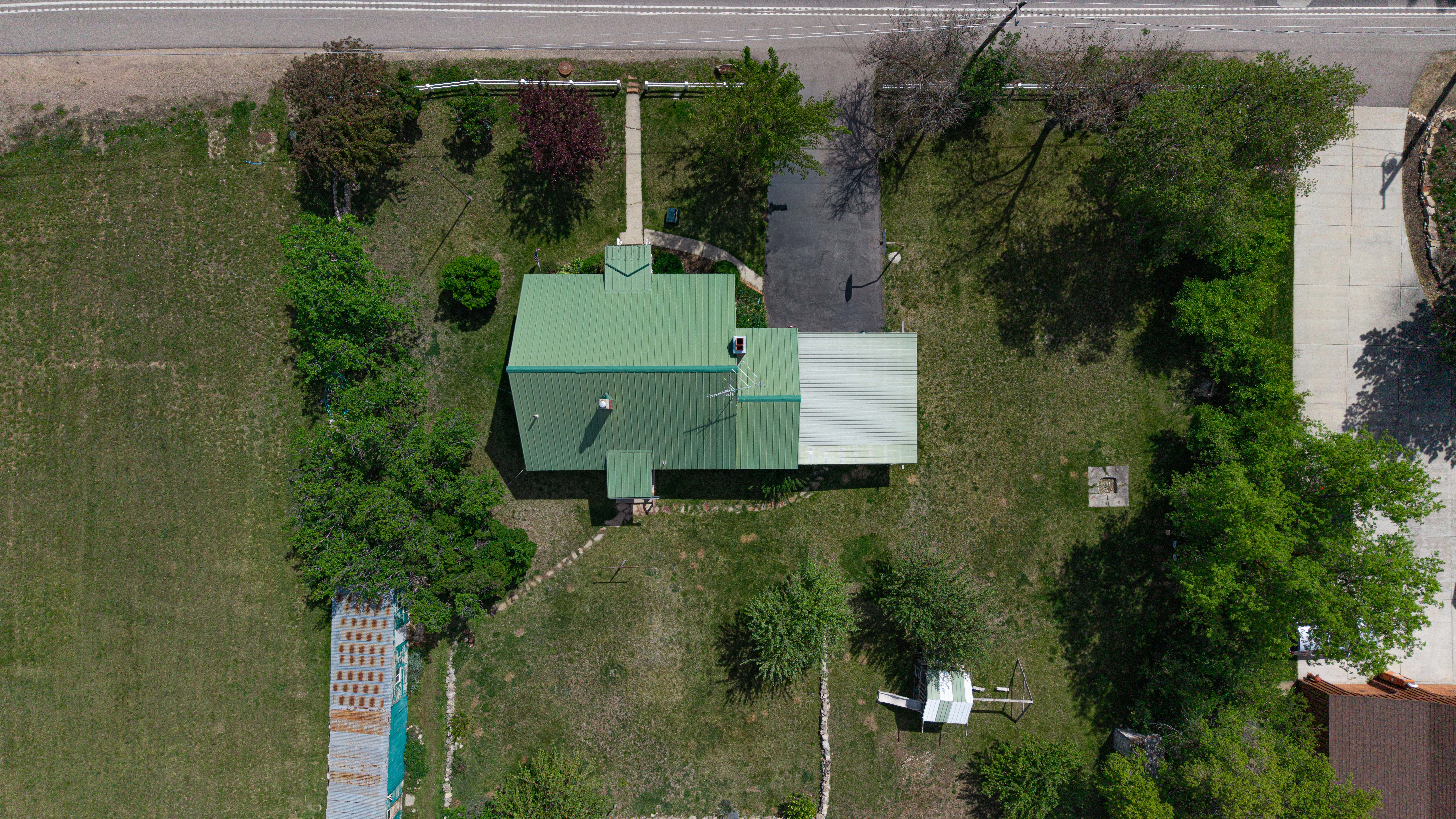 Property featuring a green metal roof, expansive green space, mature trees, and a concrete driveway