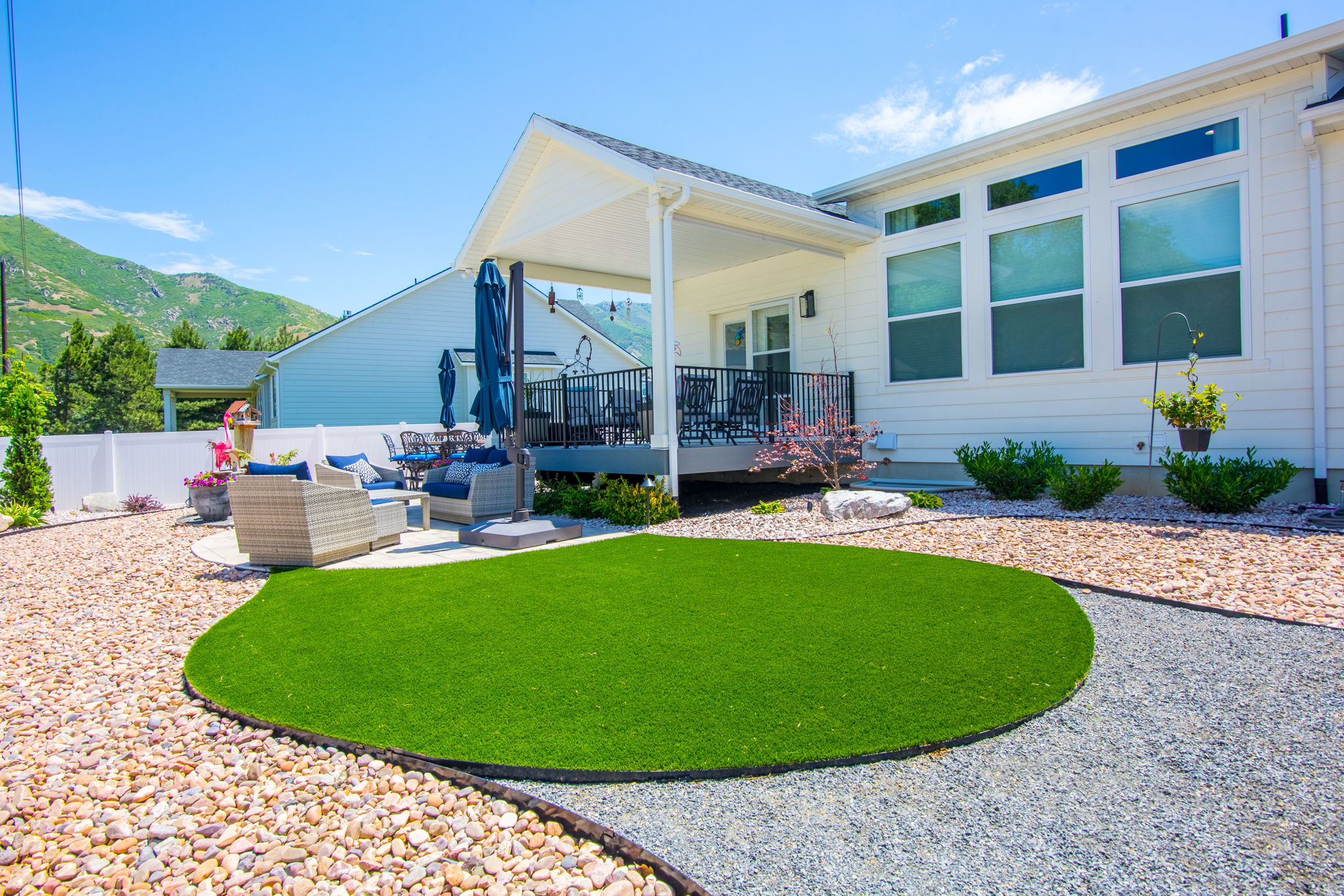 Rear exterior featuring a covered patio, composite decking, white vinyl fencing, a curved artificial turf area, and surrounding gravel landscaping