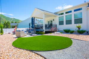 Rear exterior featuring a covered patio, composite decking, white vinyl fencing, a curved artificial turf area, and surrounding gravel landscaping