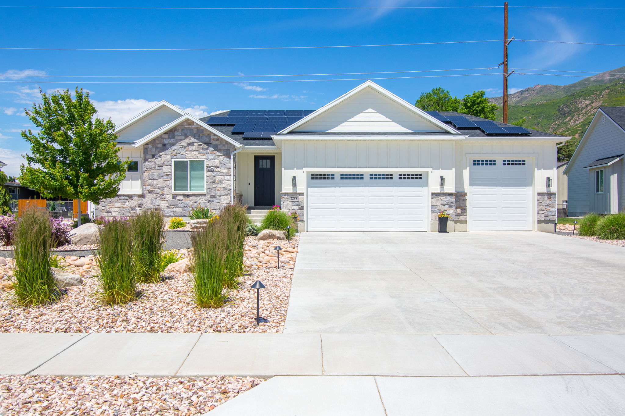Contemporary ranch-style residence featuring a stone and board-and-batten facade, solar panels, an attached three-car garage with carriage-style doors, a concrete driveway, and low-maintenance landscaping with decorative rocks and plantings