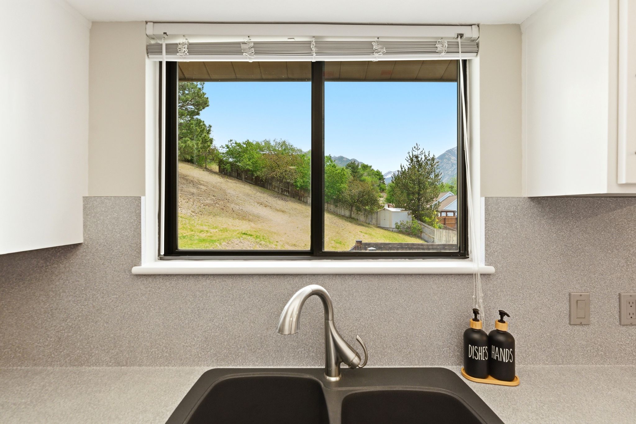 Kitchen sink with a dual basin, a high-arc faucet, and a window overlooking a sloped, treed landscape