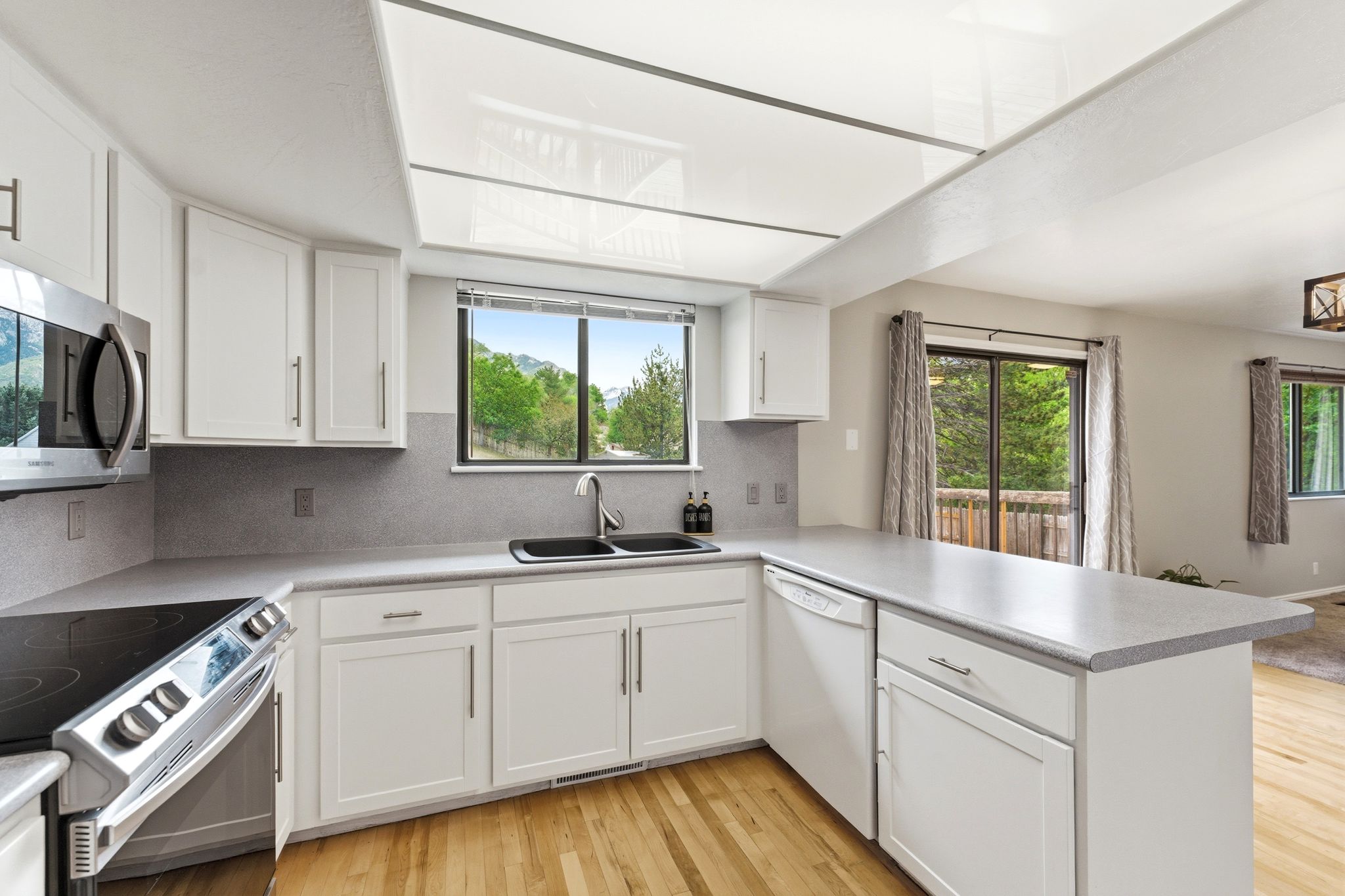 Bright kitchen featuring white cabinetry, light countertops, stainless steel appliances, wood-finish flooring, and a large window overlooking natural scenery