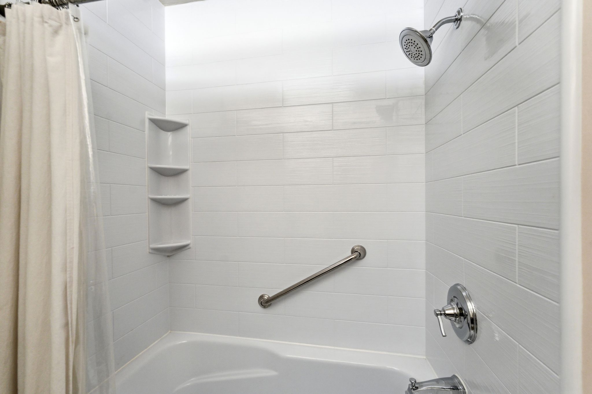 Bathtub and shower combination featuring white horizontal subway tile, chrome showerhead, and a grab bar